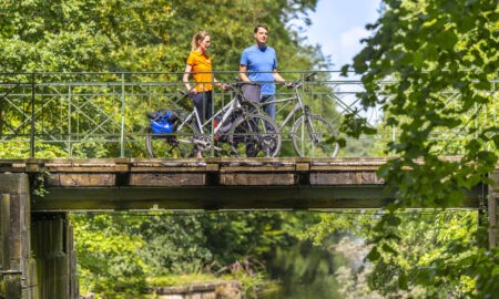 Zwei Personen stehen mit ihren Fahrrädern auf einer schmalen Metallbrücke über einem ruhigen Kanal. Die Brücke ist von dichtem Grün umgeben, während sich das Wasser unterhalb zwischen Bäumen und Sträuchern hindurchzieht.