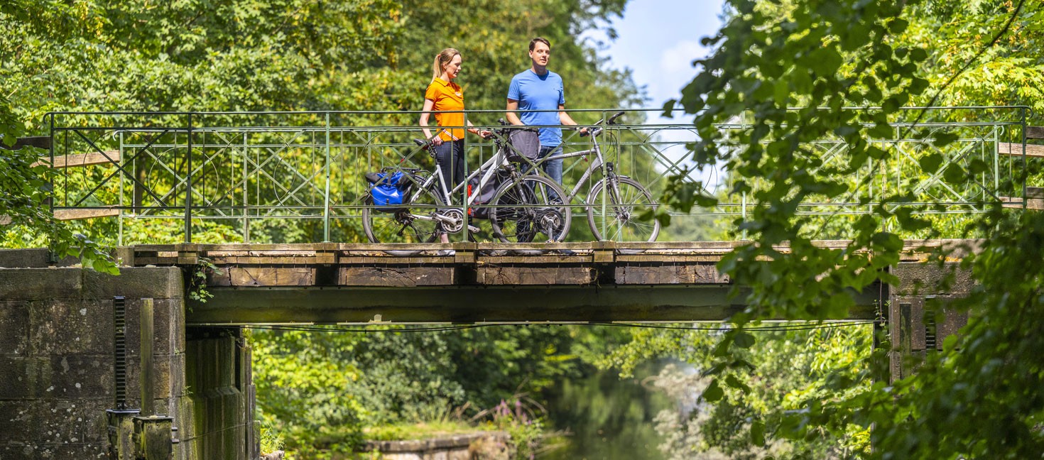 Zwei Personen stehen mit ihren Fahrrädern auf einer schmalen Metallbrücke über einem ruhigen Kanal. Die Brücke ist von dichtem Grün umgeben, während sich das Wasser unterhalb zwischen Bäumen und Sträuchern hindurchzieht.
