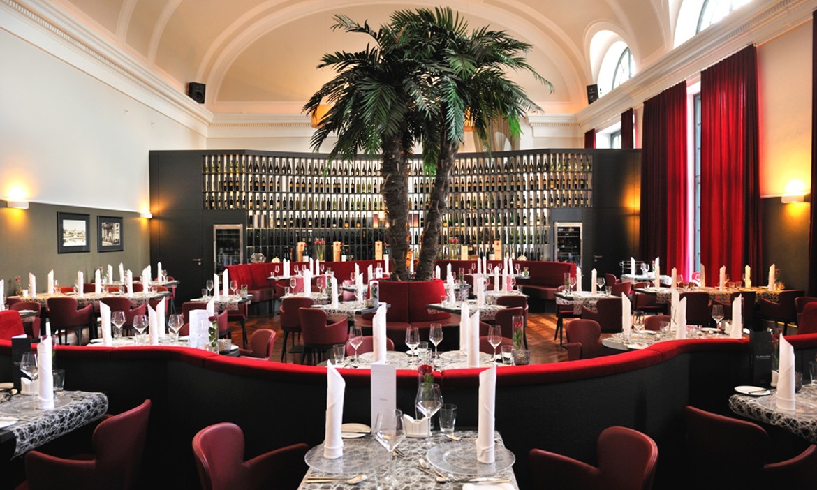 Die Innenansicht des Restaurants "Palmengarten". Inmittel steht eine Palme im Hintergrund ist eine gut befüllte Bar zu sehen.