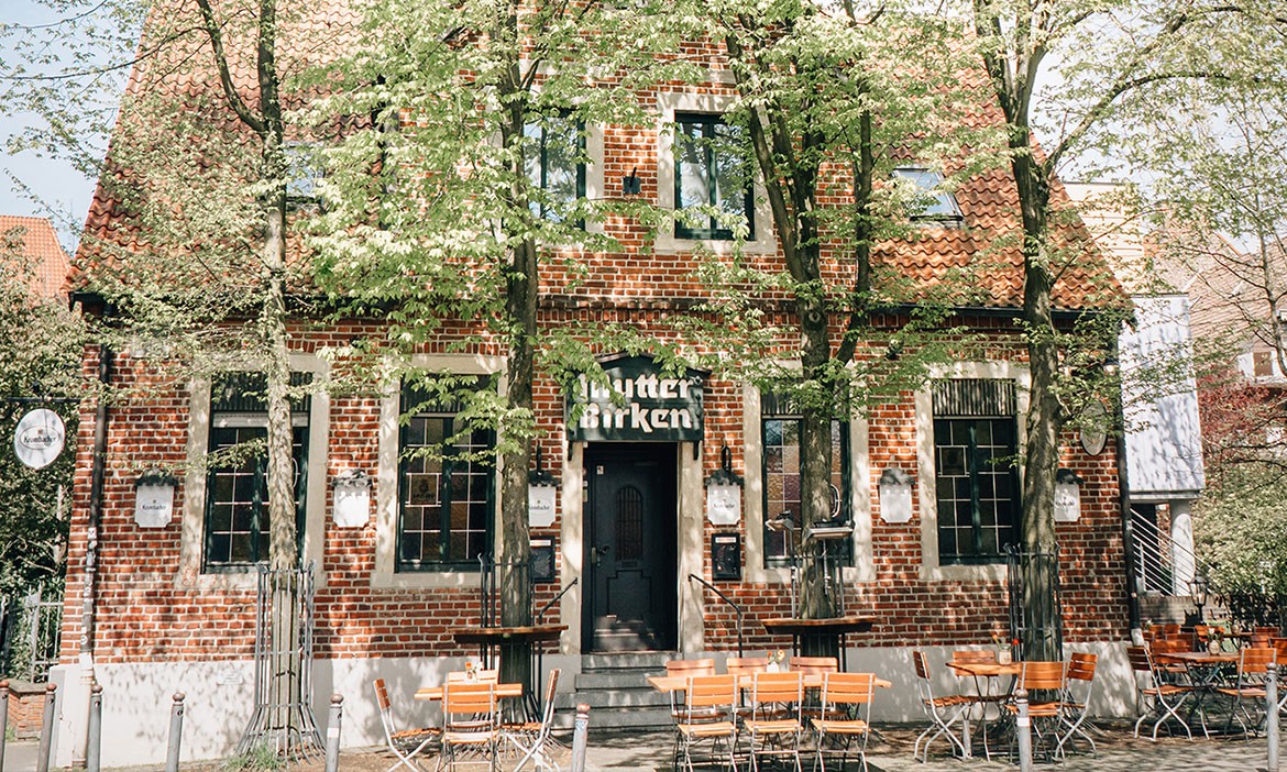 Eine einladende Außenansicht des Biergartens bei Mutter Birken im Kreuzviertel von Münster. Das historische Backsteingebäude ist von Kirschbäumen umgeben, und die Tische und Stühle des Biergartens sind bereit für Gäste.