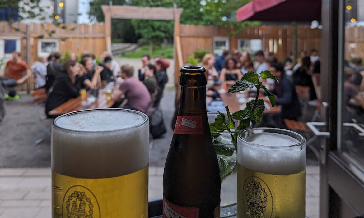 Ein lebhafter Biergarten der Roten Lola in Münster. Im Vordergrund stehen zwei Biergläser und eine Bierflasche auf einem Tisch, im Hintergrund sind zahlreiche Menschen zu sehen, die an Holzbänken sitzen und sich unterhalten. Der Biergarten ist von einem Holzzaun umgeben, und im Hintergrund sind grüne Bäume und ein Torbogen sichtbar.