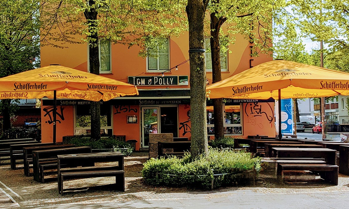Ein gemütlicher Biergarten des Lokals Tom & Polly in Münster. Große, orangefarbene Sonnenschirme spenden Schatten über den Holzbänken und Tischen. Das Gebäude im Hintergrund ist in einem warmen Orangeton gestrichen und von Bäumen umgeben, die für eine angenehme Atmosphäre sorgen.