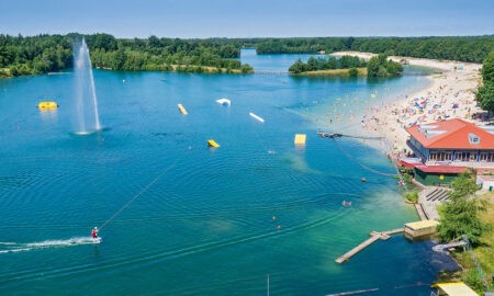 Ein idyllischer See mit klarem, blauem Wasser, umgeben von üppigem Grün. Am Ufer befindet sich ein Sandstrand mit zahlreichen Menschen, die das sonnige Wetter genießen. Es gibt eine große Wasserfontäne in der Mitte des Sees und mehrere aufblasbare Wasserhindernisse. Ein Wakeboarder fährt über das Wasser, gezogen von einem Seil. Am rechten Ufer steht ein Gebäude mit rotem Dach, vermutlich ein Restaurant oder Café, und weitere Einrichtungen für Besucher. Der Himmel ist klar und blau, was auf einen sonnigen Tag hinweist.