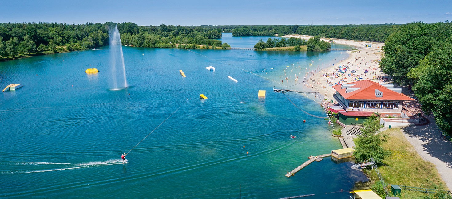 Ein idyllischer See mit klarem, blauem Wasser, umgeben von üppigem Grün. Am Ufer befindet sich ein Sandstrand mit zahlreichen Menschen, die das sonnige Wetter genießen. Es gibt eine große Wasserfontäne in der Mitte des Sees und mehrere aufblasbare Wasserhindernisse. Ein Wakeboarder fährt über das Wasser, gezogen von einem Seil. Am rechten Ufer steht ein Gebäude mit rotem Dach, vermutlich ein Restaurant oder Café, und weitere Einrichtungen für Besucher. Der Himmel ist klar und blau, was auf einen sonnigen Tag hinweist.