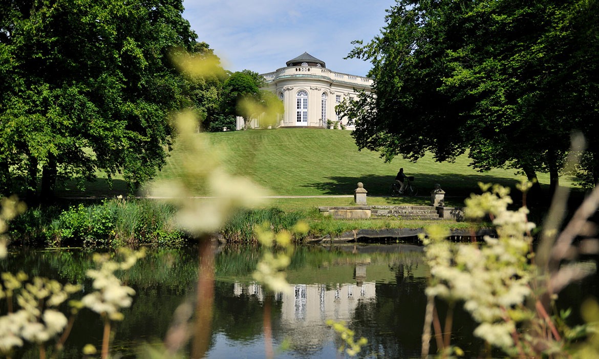 Ein malerischer Blick auf eine historische Villa, die auf einem sanften Hügel thront, umgeben von üppigem Grün. Im Vordergrund spiegelt sich die Villa im ruhigen Wasser eines Teiches, der von Blumen und Pflanzen umrahmt ist. Die Szene vermittelt eine friedliche, fast idyllische Atmosphäre, die perfekt zu einem entspannten Tag in der Natur passt. Ein Radfahrer ist im Hintergrund zu sehen, was das Bild der Ruhe und Entspannung abrundet.