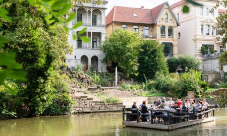 Ein Floß gleitet gemächlich die Oker entlang, vorbei an grünen Ufern und historischen Villen. Auf dem Floß sitzen Menschen, die die entspannte Atmosphäre genießen und die umliegende Natur bewundern. Im Hintergrund sind gut erhaltene Gebäude mit charmanter Architektur zu sehen, die den historischen Charakter der Umgebung unterstreichen. Die Szene wird von üppigem Grün und friedlichem Wasser umrahmt, was ein Gefühl von Ruhe und Gelassenheit vermittelt.