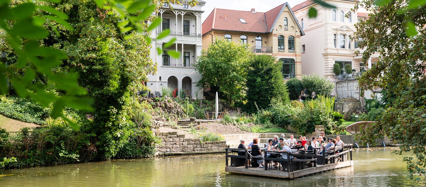 Ein Floß gleitet gemächlich die Oker entlang, vorbei an grünen Ufern und historischen Villen. Auf dem Floß sitzen Menschen, die die entspannte Atmosphäre genießen und die umliegende Natur bewundern. Im Hintergrund sind gut erhaltene Gebäude mit charmanter Architektur zu sehen, die den historischen Charakter der Umgebung unterstreichen. Die Szene wird von üppigem Grün und friedlichem Wasser umrahmt, was ein Gefühl von Ruhe und Gelassenheit vermittelt.