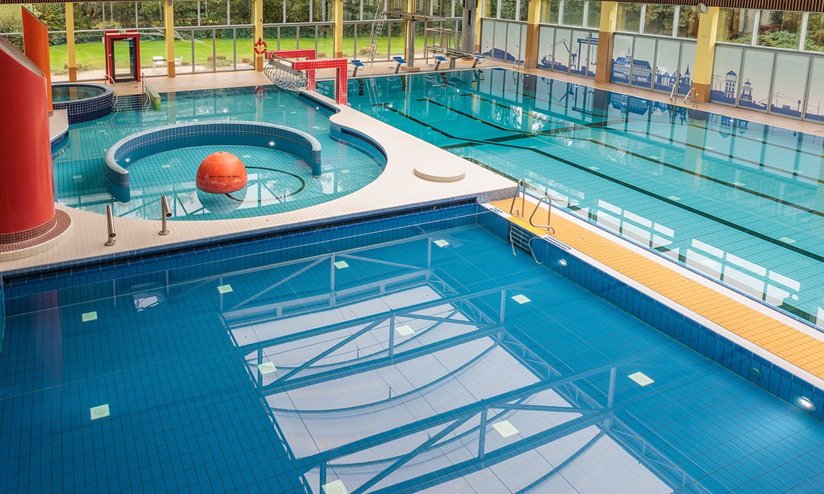 Das Bild zeigt einen modernen Innenbereich eines Schwimmbads mit mehreren Becken. Im Vordergrund ist ein großes, tiefes Schwimmbecken zu sehen, während sich in der Mitte ein kreisförmiger Pool mit einer großen orangefarbenen Kugel befindet. Im Hintergrund befindet sich ein weiteres rechteckiges Becken mit abgetrennten Bahnen zum Schwimmen. Die Becken sind von hellen Fliesen umgeben und durch große Fenster strömt viel Tageslicht herein, wodurch der Blick ins Grüne freigegeben wird.