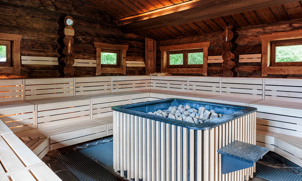 Das Bild zeigt das Innere einer traditionellen finnischen Sauna aus Holz mit hellen Sitzbänken und dunklen, rustikalen Holzwänden. In der Mitte befindet sich ein Saunaofen mit Lavasteinen. Kleine Fenster lassen natürliches Licht herein, und ein Thermometer sowie ein Hygrometer hängen an der Wand.