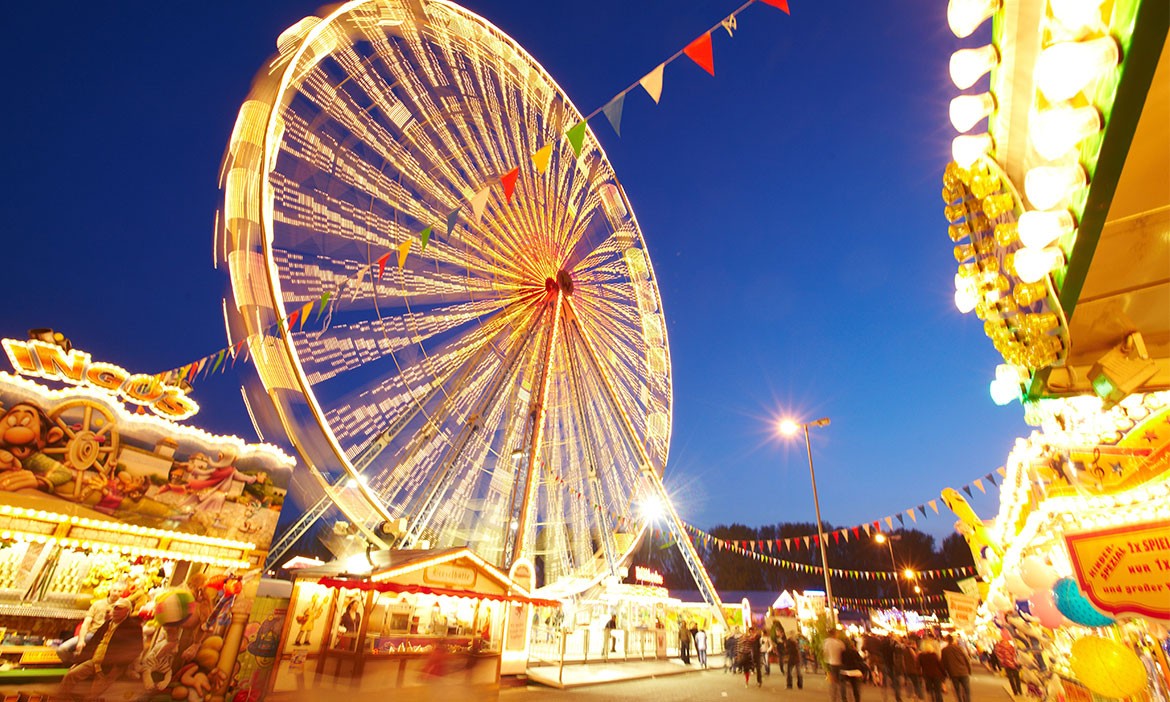 Das Bild zeigt eine fröhliche Jahrmarkt-Szene bei Nacht mit einem großen, beleuchteten Riesenrad im Zentrum, das in gelben und weißen Lichtern erstrahlt. Bunte Wimpelketten hängen quer durch das Bild und verstärken die festliche Atmosphäre. Im Vordergrund sind beleuchtete Verkaufsstände mit farbenfrohen Schildern und Dekorationen zu sehen, die typische Jahrmarkt-Attraktionen wie Spiele und Snacks anbieten. Der Himmel ist tiefblau, was die Lichter des Riesenrads und der Stände noch intensiver leuchten lässt. Die Szenerie ist belebt, mit Besuchern, die den Rummelplatz genießen.