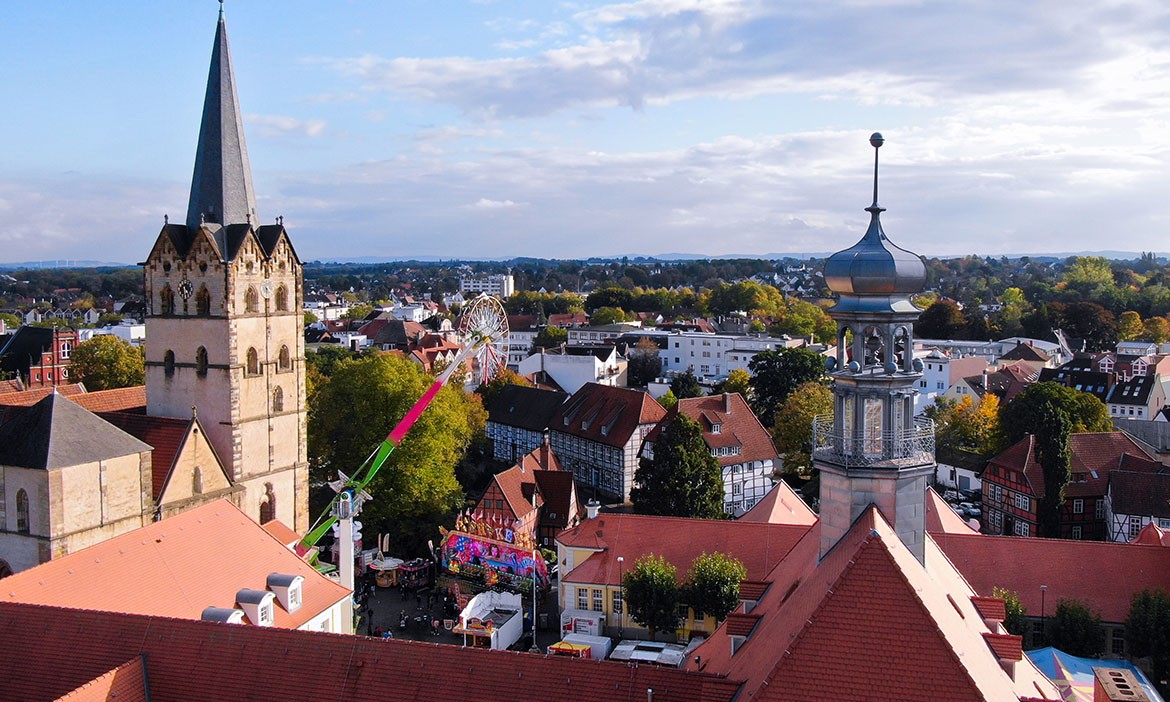 Das Bild zeigt eine Vogelperspektive auf das Stadtzentrum von Herford mit historischen Gebäuden, darunter ein markanter Kirchturm mit Spitze und ein weiteres Gebäude mit einem barocken Zwiebelturm. Im Hintergrund ist ein Riesenrad sowie ein Fahrgeschäft mit leuchtenden Farben sichtbar, die Teil eines Rummels sind. Die Dächer der Fachwerkhäuser sind mit roten Ziegeln bedeckt, und der Herbst ist an den bunt gefärbten Bäumen in der Stadtlandschaft erkennbar. Der Himmel ist teilweise bewölkt, was der Szenerie einen malerischen, lebendigen Charakter verleiht.