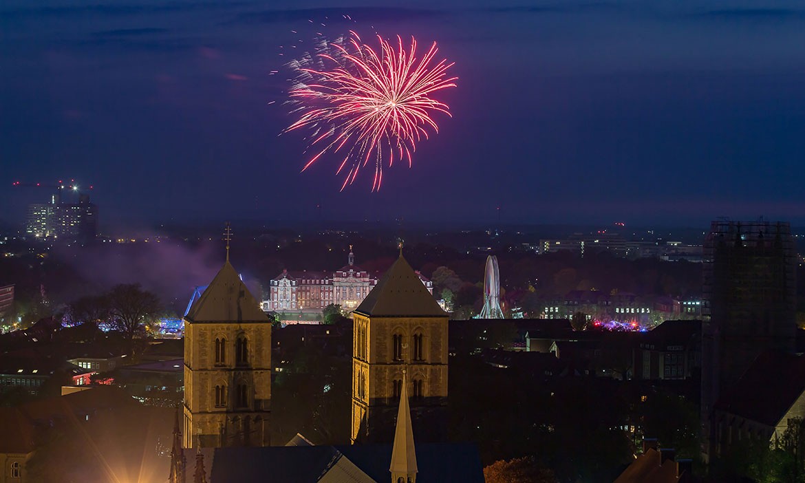 
Das Bild zeigt ein nächtliches Feuerwerk. Im Vordergrund sind die beleuchteten Türme eines historischen Gebäudes zu sehen. Im Hintergrund erhellt ein rotes Feuerwerk den dunkelblauen Nachthimmel und zieht die Blicke auf sich. Unten am Bildrand sind beleuchtete Fahrgeschäfte, darunter ein großes Riesenrad, sowie eine festlich geschmückte Veranstaltung zu erkennen. Die Szene vermittelt eine festliche und magische Atmosphäre, während die Lichter der Stadt und der Veranstaltung den Abend erleuchten.