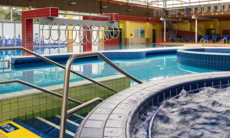 Das Bild zeigt ein modernes Hallenbad mit mehreren Pools. Im Vordergrund befindet sich ein Whirlpool mit sprudelndem Wasser. Eine Edelstahl-Treppenstange führt in den angrenzenden Schwimmbereich mit mehreren Becken unterschiedlicher Tiefe. An der Seite ist ein Spielbereich mit Ringen, die über das Wasser gespannt sind. Die Fliesen der Becken sind in Blau- und Grüntönen gehalten, während die Wände des Gebäudes in warmen Gelb- und Rottönen gestaltet sind. Im Hintergrund gibt es Liegestühle auf einer erhöhten Plattform.