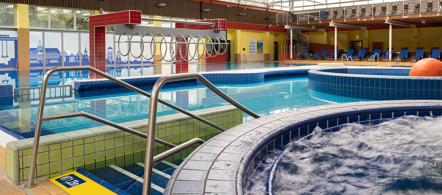 Das Bild zeigt ein modernes Hallenbad mit mehreren Pools. Im Vordergrund befindet sich ein Whirlpool mit sprudelndem Wasser. Eine Edelstahl-Treppenstange führt in den angrenzenden Schwimmbereich mit mehreren Becken unterschiedlicher Tiefe. An der Seite ist ein Spielbereich mit Ringen, die über das Wasser gespannt sind. Die Fliesen der Becken sind in Blau- und Grüntönen gehalten, während die Wände des Gebäudes in warmen Gelb- und Rottönen gestaltet sind. Im Hintergrund gibt es Liegestühle auf einer erhöhten Plattform.