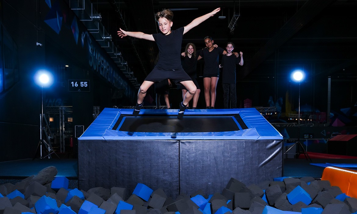 Das Bild zeigt ein Kind, das auf einem Trampolin in einer Indoor-Sporthalle springt. Im Vordergrund befindet sich ein Schaumstoffbecken, das mit blauen und grauen Schaumstoffwürfeln gefüllt ist. Im Hintergrund stehen drei weitere Kinder, die dem Trampolinspringer zuschauen. Die Halle ist dunkel, aber gut beleuchtet mit mehreren hellen Scheinwerfern. Alle Kinder tragen schwarze Sportkleidung.