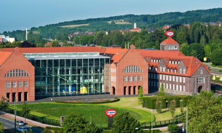 Das Bild zeigt eine Luftaufnahme des Dr. Oetker Hauptsitzes in Bielefeld. Das markante Gebäude besteht aus einer Kombination von historischem roten Backsteinbau und modernen architektonischen Elementen. In der Mitte befindet sich ein großer, verglaster Bereich, der die beiden älteren Gebäudeflügel miteinander verbindet und viel Licht ins Innere lässt. Durch die Glasfassade ist das auffällige gelbe Objekt, das in den vorherigen Bildern zu sehen war, erkennbar. Das Gebäude ist von einer gepflegten Grünanlage umgeben, und im Hintergrund erstreckt sich eine hügelige Landschaft. Mehrere Dr. Oetker Logos auf dem Gelände weisen auf den Unternehmenssitz hin.