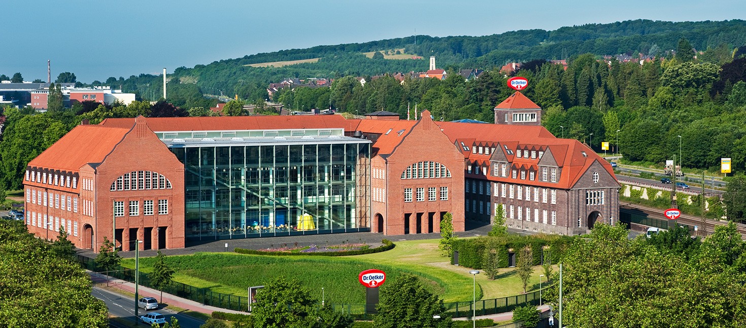 Das Bild zeigt eine Luftaufnahme des Dr. Oetker Hauptsitzes in Bielefeld. Das markante Gebäude besteht aus einer Kombination von historischem roten Backsteinbau und modernen architektonischen Elementen. In der Mitte befindet sich ein großer, verglaster Bereich, der die beiden älteren Gebäudeflügel miteinander verbindet und viel Licht ins Innere lässt. Durch die Glasfassade ist das auffällige gelbe Objekt, das in den vorherigen Bildern zu sehen war, erkennbar. Das Gebäude ist von einer gepflegten Grünanlage umgeben, und im Hintergrund erstreckt sich eine hügelige Landschaft. Mehrere Dr. Oetker Logos auf dem Gelände weisen auf den Unternehmenssitz hin.