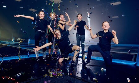 Das Bild zeigt eine Gruppe von sechs Kindern, die auf einer Indoor-Trampolinanlage feiern. Sie sind von bunten Luftschlangen und Konfetti umgeben, die in der Luft schweben. Alle Kinder tragen sportliche, schwarze Kleidung und wirken fröhlich und ausgelassen. Einige Kinder posieren auf dem Trampolin, während andere in die Kamera lächeln und den Moment genießen. Die Szene ist lebendig und festlich, mit bunten Lichtern und Konfetti, das die fröhliche Stimmung unterstreicht.