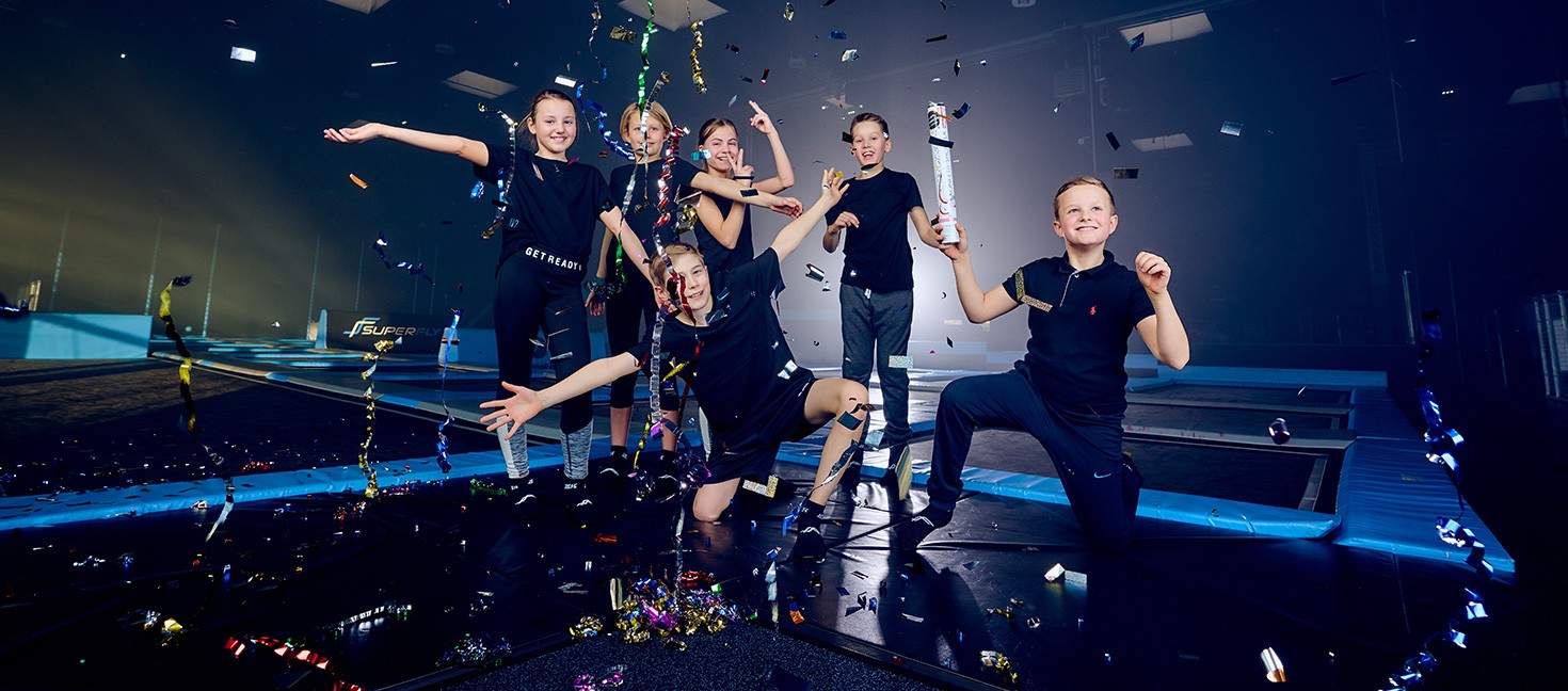 Das Bild zeigt eine Gruppe von sechs Kindern, die auf einer Indoor-Trampolinanlage feiern. Sie sind von bunten Luftschlangen und Konfetti umgeben, die in der Luft schweben. Alle Kinder tragen sportliche, schwarze Kleidung und wirken fröhlich und ausgelassen. Einige Kinder posieren auf dem Trampolin, während andere in die Kamera lächeln und den Moment genießen. Die Szene ist lebendig und festlich, mit bunten Lichtern und Konfetti, das die fröhliche Stimmung unterstreicht.