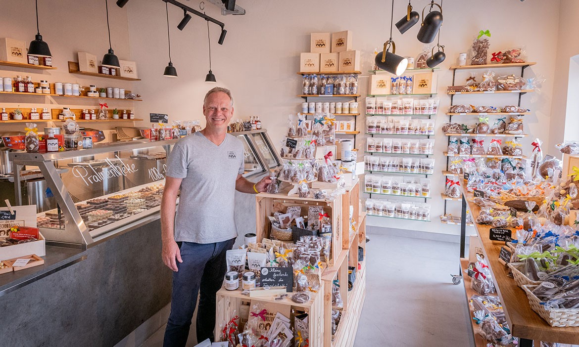 Ein freundlicher Mann steht im hellen, einladenden Geschäft der drei Schokojungs mit einer großen Auswahl an verpackten Süßigkeiten, Pralinen und Feinkostprodukten. Holzregale und Vitrinen präsentieren die Produkte, während dekorative Beleuchtung den Raum warm und einladend wirken lässt.