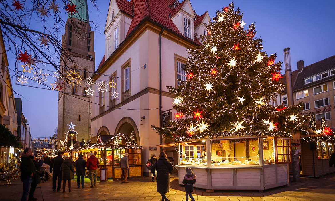 Dieses Bild zeigt einen Weihnachtsmarkt in einer malerischen europäischen Altstadt. Im Mittelpunkt steht ein prächtig geschmückter Weihnachtsbaum, der mit leuchtenden Sternen in Rot und Weiß dekoriert ist. Umgeben ist der Baum von charmanten Holzhütten, in denen Waren und Leckereien angeboten werden. Im Hintergrund erhebt sich ein historischer Kirchturm, und Lichterketten mit Sternmotiven hängen über den Köpfen der Besucher, die die festliche Atmosphäre genießen. Die Dämmerung sorgt für eine stimmungsvolle Beleuchtung, die den Weihnachtszauber unterstreicht.