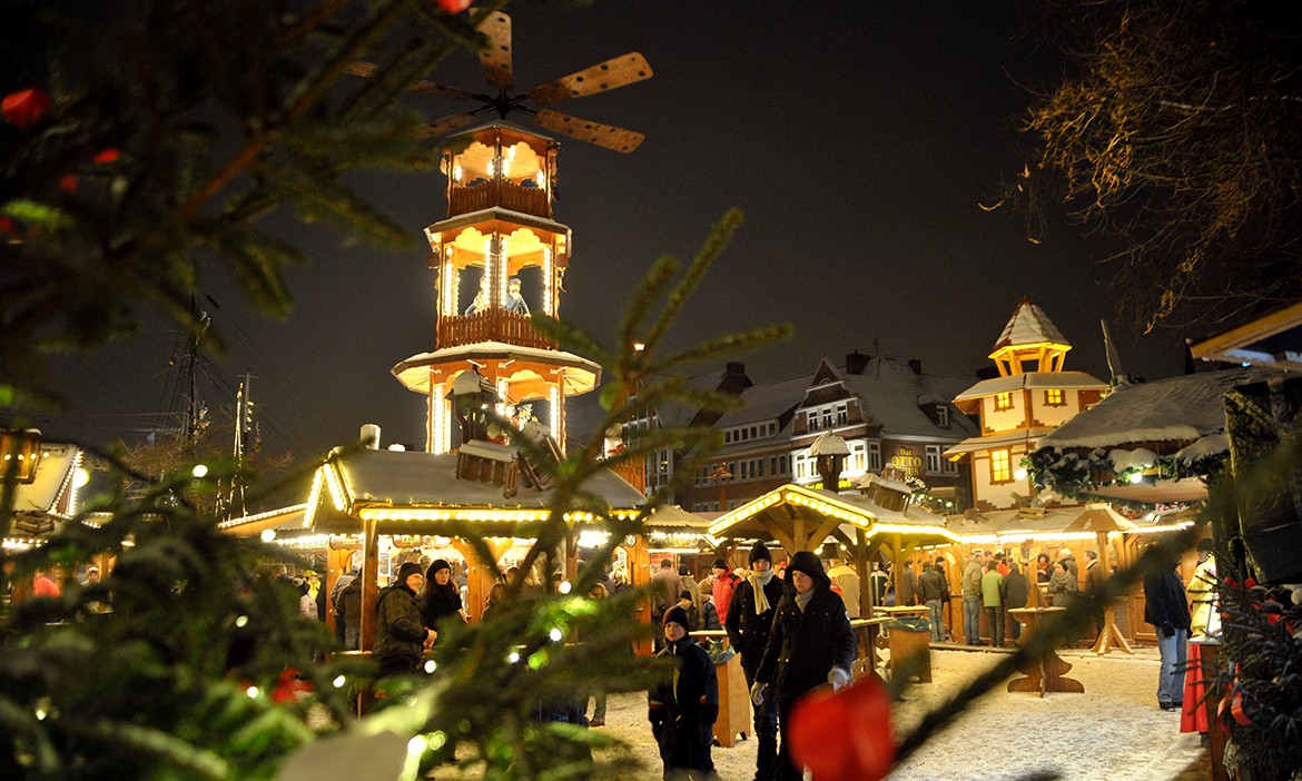 Dieses Bild zeigt einen idyllischen Weihnachtsmarkt bei Nacht, mit verschneitem Boden und warm beleuchteten Holzhütten. Im Zentrum steht eine traditionelle, festlich dekorierte Weihnachtspyramide mit mehreren Etagen, die mit Lichtern und Figuren geschmückt ist. Besucher, in warme Winterkleidung gehüllt, spazieren durch den Markt, während die Atmosphäre von funkelnden Lichtern und winterlicher Romantik erfüllt ist. Im Vordergrund sind Tannenzweige mit roten Dekorationen zu sehen, die das Bild einrahmen. Die verschneiten Dächer der umliegenden Gebäude verleihen der Szene zusätzlichen Charme.