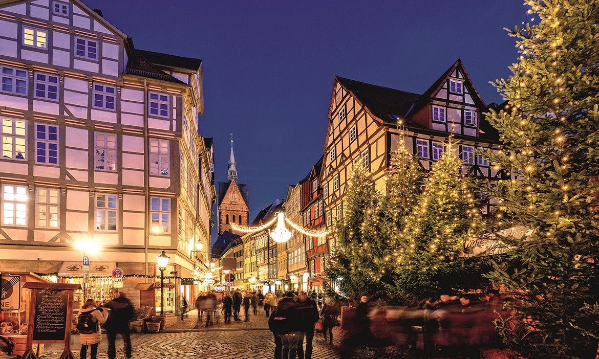 Dieses Bild zeigt einen Weihnachtsmarkt in einer malerischen Fachwerkstadt. Die historische Architektur der Fachwerkhäuser wird durch warme Lichter beleuchtet, während Girlanden und ein funkelnder Lichterbogen über die Straße gespannt sind. Im Vordergrund stehen festlich geschmückte Tannenbäume mit leuchtenden Lichtern, die den Marktbesuchern eine gemütliche Atmosphäre bieten. Im Hintergrund ragt der Kirchturm in den Nachthimmel, der die Szenerie mit seinem Charme ergänzt. Menschen flanieren durch die Kopfsteinpflasterstraßen und genießen die weihnachtliche Stimmung.