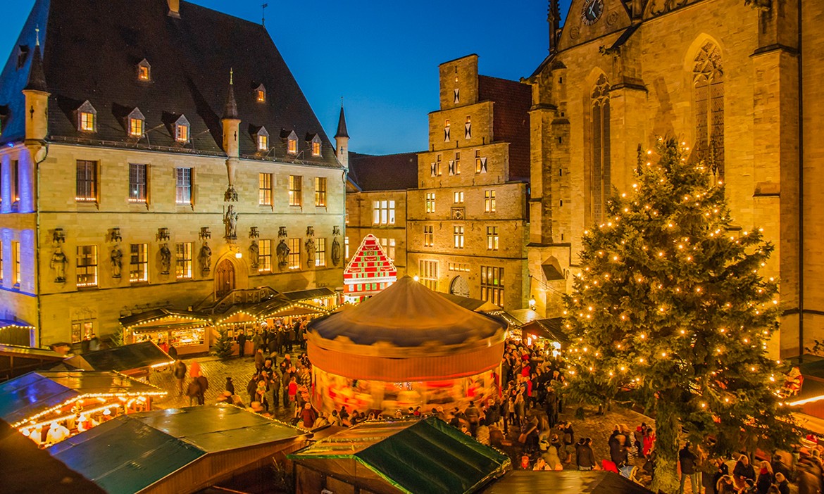 Dieses Bild zeigt eine festliche Weihnachtsmarktszene in einem historischen europäischen Stadtkern. Der Markt ist mit warmen Lichtern, bunten Ständen und einem großen, festlich beleuchteten Weihnachtsbaum auf der rechten Seite geschmückt. Im Zentrum des Geschehens steht ein Karussell, umgeben von Besuchern, die die festliche Stimmung genießen. Die historischen Gebäude mit beleuchteten Fenstern im Hintergrund verstärken die gemütliche und einladende Atmosphäre in der Dämmerung.