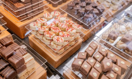 Eine Nahaufnahme von verschiedenen Pralinen, die sorgfältig auf Holztabletts in einer Vitrine präsentiert werden. Es gibt eine Vielfalt an Schokoladen in unterschiedlichen Formen, Farben und Verzierungen, darunter weiße Pralinen mit roten Streuseln, Vollmilchpralinen mit Mustern und dunkle Schokoladenwürfel. Die Präsentation wirkt hochwertig und appetitlich.