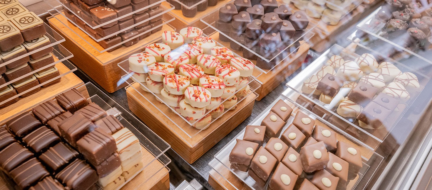 Eine Nahaufnahme von verschiedenen Pralinen, die sorgfältig auf Holztabletts in einer Vitrine präsentiert werden. Es gibt eine Vielfalt an Schokoladen in unterschiedlichen Formen, Farben und Verzierungen, darunter weiße Pralinen mit roten Streuseln, Vollmilchpralinen mit Mustern und dunkle Schokoladenwürfel. Die Präsentation wirkt hochwertig und appetitlich.