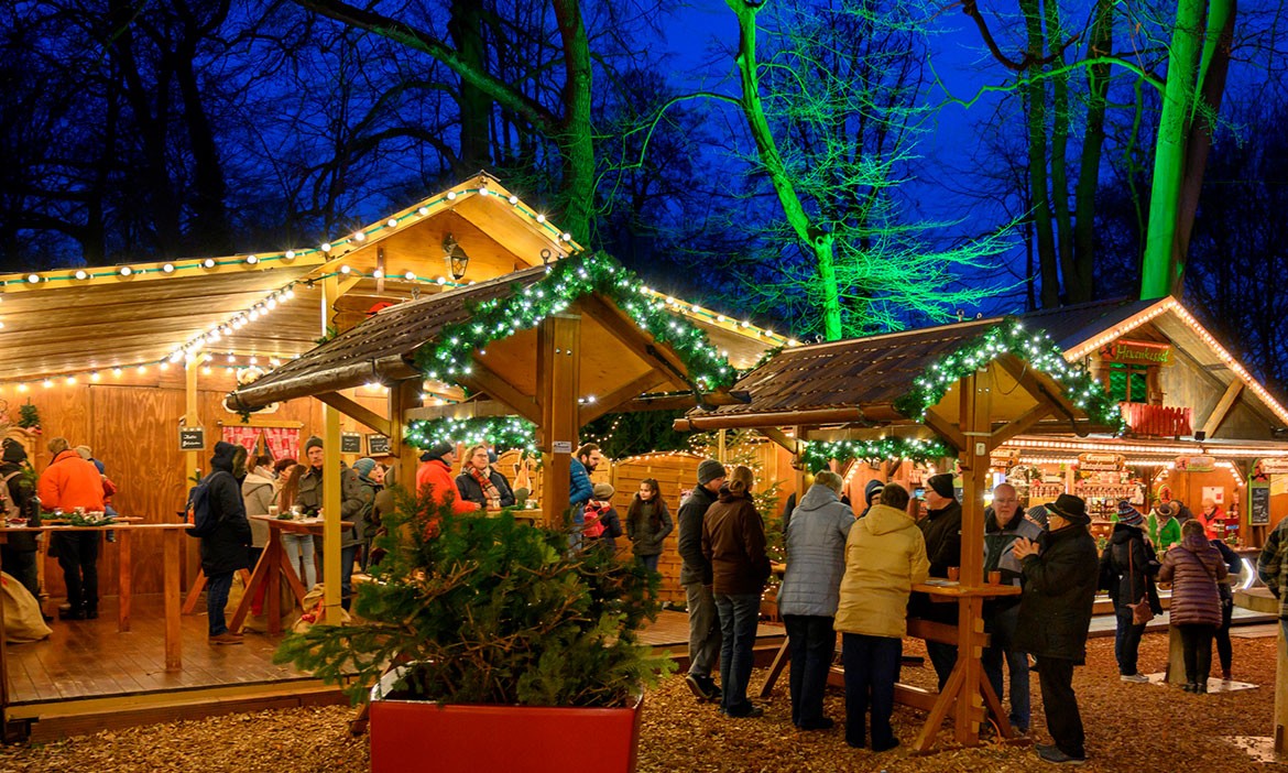 Ein festlicher Weihnachtsmarkt bei Dämmerung: Holzhütten mit Lichterketten und grünen Girlanden beleuchtet. Menschen in Winterkleidung genießen Speisen und Getränke an Stehtischen. Im Hintergrund sind beleuchtete Bäume mit grünem Licht und ein dunkler Abendhimmel zu sehen.