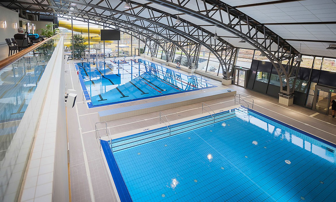 Modernes Hallenbad mit großen Schwimmbecken und einer markanten Stahlkonstruktion. Im Vordergrund ein Nichtschwimmerbereich, dahinter ein Wettkampfbecken mit mehreren Bahnen. Eine gelbe Wasserrutsche ist im Hintergrund zu sehen. Die großzügigen Fensterfronten lassen viel Tageslicht herein. 