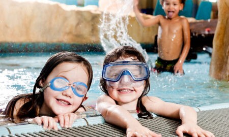 Zwei lachende Kinder mit Taucherbrillen genießen das Wasser im Erlebnisbad, während im Hintergrund ein Junge spielerisch Wasser verspritzt. Die Szene vermittelt Freude und unbeschwerten Badespaß in einer familienfreundlichen Umgebung.