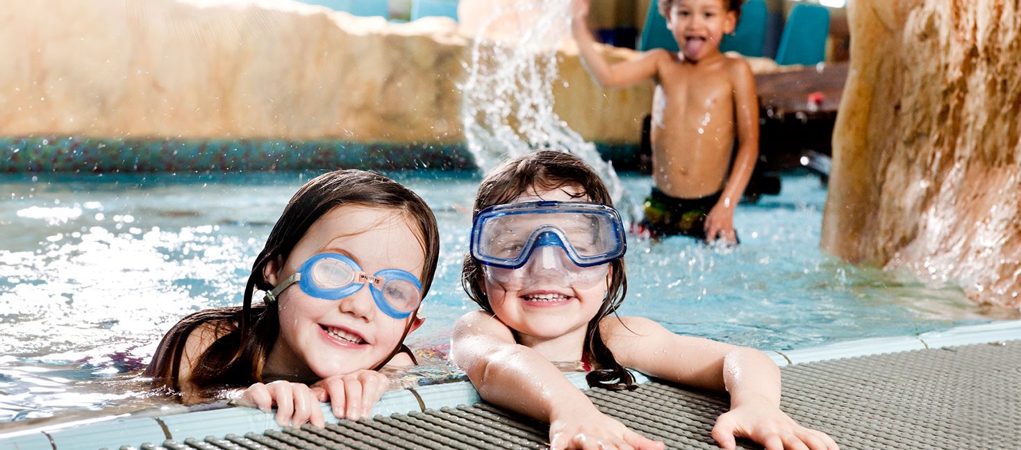 Zwei lachende Kinder mit Taucherbrillen genießen das Wasser im Erlebnisbad, während im Hintergrund ein Junge spielerisch Wasser verspritzt. Die Szene vermittelt Freude und unbeschwerten Badespaß in einer familienfreundlichen Umgebung.