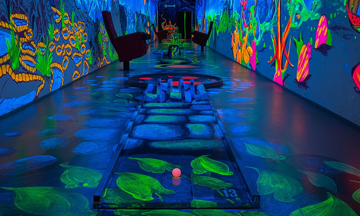 Das Bild zeigt eine Bahn eines Schwarzlicht-Minigolf-Parcours mit fluoreszierenden Farben und fantasievoller Gestaltung. Der Boden ist mit leuchtenden Mustern bemalt, darunter grüne Blätter und eine illusionäre Steinbrücke. Eine pinkfarbene Minigolfkugel liegt am Start der Bahn, die mit Hindernissen versehen ist. Die Wände sind mit farbenfrohen, surrealen Motiven bemalt, die unter UV-Licht intensiv leuchten. Im Hintergrund sind Sitzgelegenheiten und weitere Bahnen zu sehen, die in das immersive, leuchtende Design integriert sind.