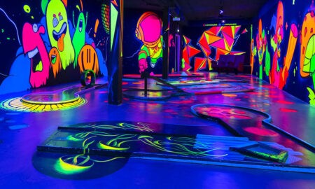 Das Bild zeigt eine Schwarzlicht-Minigolfanlage mit mehreren Bahnen, die durch leuchtende Farben und fantasievolle Wandmalereien eine immersive Atmosphäre schaffen. Die Wände sind mit fluoreszierenden Motiven wie bunten Geistern, einem Astronauten, riesigen Pilzen und geometrischen Mustern verziert. Der Boden und die Minigolfbahnen selbst sind ebenfalls mit leuchtenden Elementen gestaltet, darunter geschwungene Linien, Kreise und Hindernisse. Das UV-Licht verstärkt die kräftigen Farben und sorgt für ein einzigartiges Spielerlebnis in einer surreal wirkenden Umgebung.