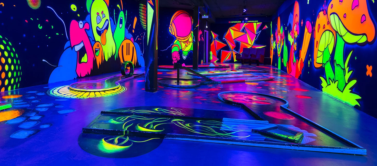 Das Bild zeigt eine Schwarzlicht-Minigolfanlage mit mehreren Bahnen, die durch leuchtende Farben und fantasievolle Wandmalereien eine immersive Atmosphäre schaffen. Die Wände sind mit fluoreszierenden Motiven wie bunten Geistern, einem Astronauten, riesigen Pilzen und geometrischen Mustern verziert. Der Boden und die Minigolfbahnen selbst sind ebenfalls mit leuchtenden Elementen gestaltet, darunter geschwungene Linien, Kreise und Hindernisse. Das UV-Licht verstärkt die kräftigen Farben und sorgt für ein einzigartiges Spielerlebnis in einer surreal wirkenden Umgebung.