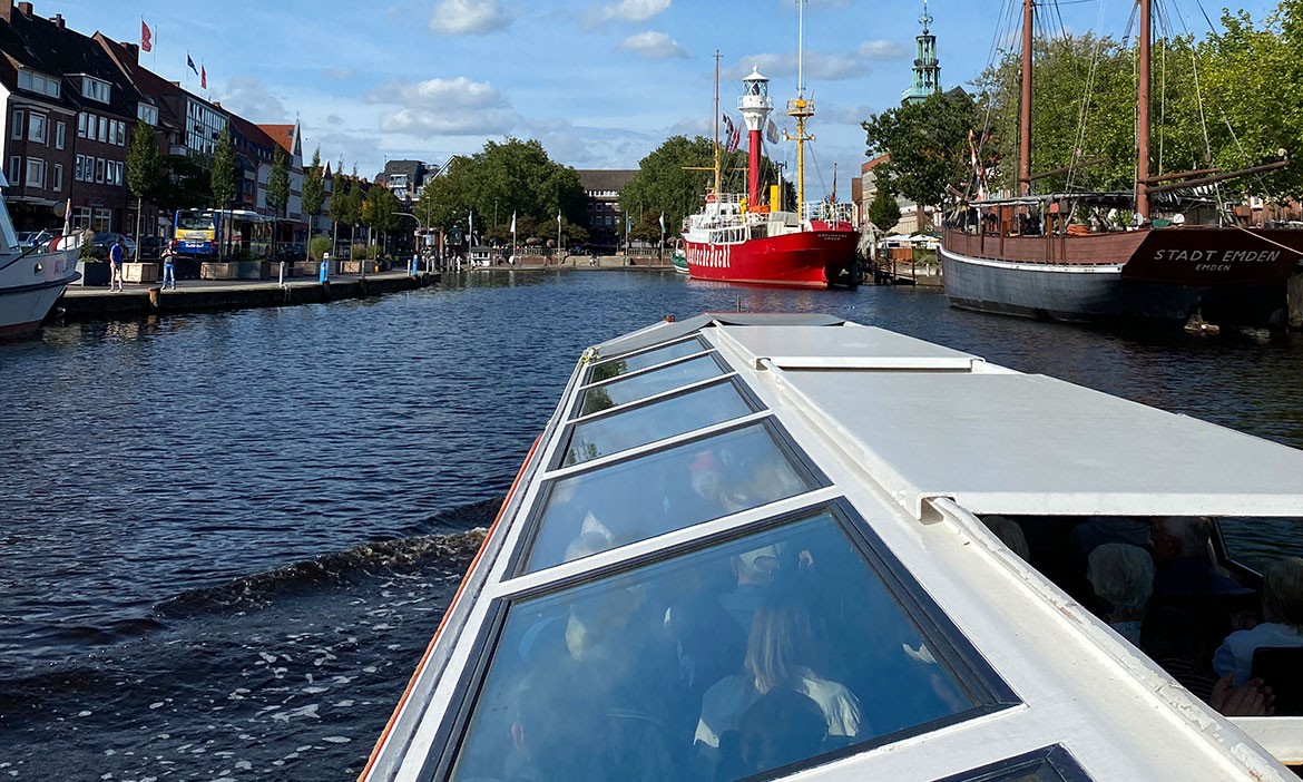 Ein Ausflugsboot fährt auf einem breiten Kanal durch Emden. Vorne sieht man das Glasdach des Bootes und die Menschen darunter. Rechts im Wasser liegt ein großes rotes Schiff mit einem weißen Turm – das ist das Feuerschiff „Amrumbank“. Daneben liegt ein altes Holzsegelboot mit dem Namen „Stadt Emden“. Links am Ufer stehen rote Häuser mit kleinen Flaggen und Bäumen. Am Himmel sind ein paar weiße Wolken.