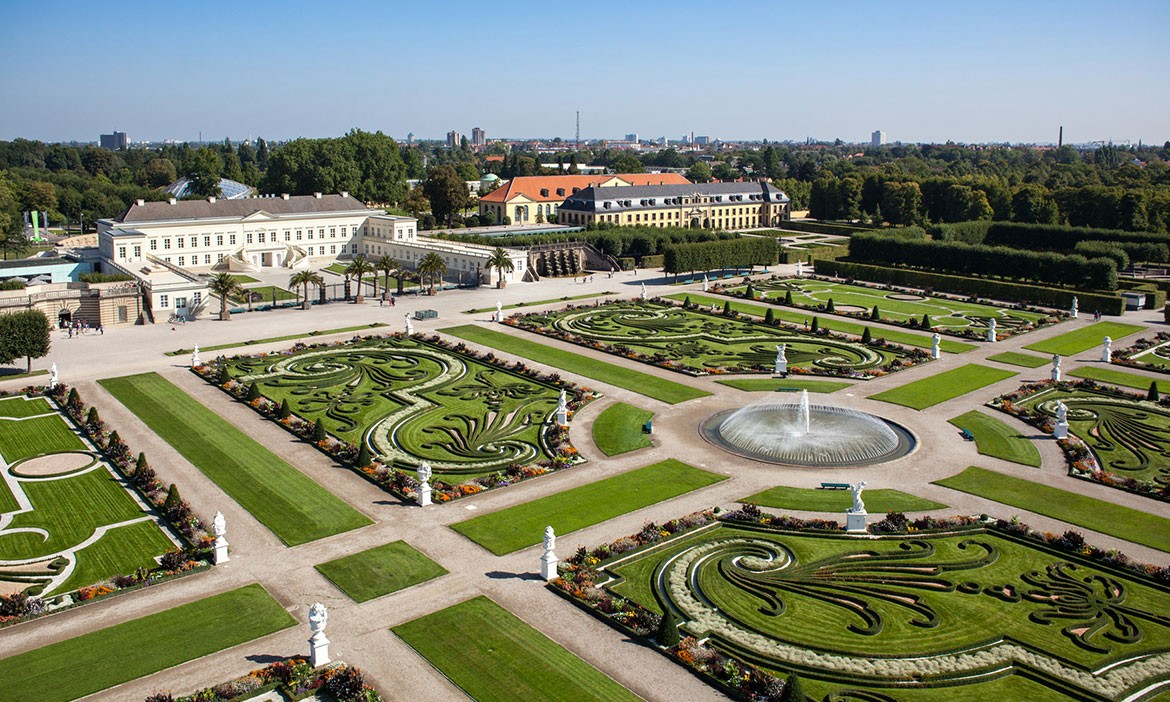 Panoramablick auf den Großen Garten der Herrenhäuser Gärten in Hannover aus der Vogelperspektive: Im Zentrum eine runde Wasserfontäne, umgeben von kunstvoll geschnittenen Buchsornamenten in barocker Symmetrie. Rechts und links rahmen akkurate Hecken und Skulpturen die Gartenarchitektur ein. Im Hintergrund das weiße Galeriegebäude und das Schloss, dahinter die Skyline von Hannover.