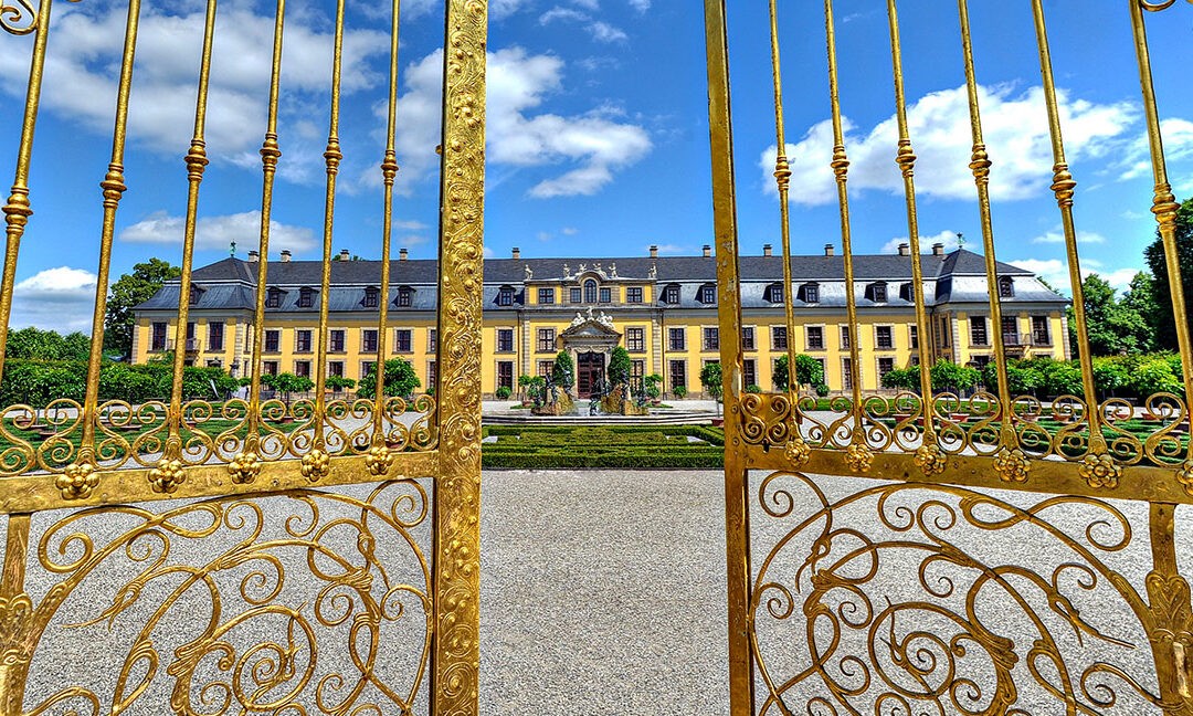 Blick durch ein prunkvoll verziertes, goldenes Eisengittertor auf das barocke Schloss Herrenhausen in Hannover. Im Vordergrund das geöffnete Tor mit floralen Ornamenten, im Hintergrund die symmetrische Gartenanlage mit Brunnen und gepflegten Beeten. Der Himmel ist strahlend blau mit einzelnen weißen Wolken – ein sonniger, klarer Tag.