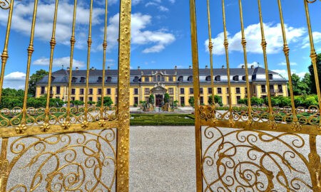 Blick durch ein prunkvoll verziertes, goldenes Eisengittertor auf das barocke Schloss Herrenhausen in Hannover. Im Vordergrund das geöffnete Tor mit floralen Ornamenten, im Hintergrund die symmetrische Gartenanlage mit Brunnen und gepflegten Beeten. Der Himmel ist strahlend blau mit einzelnen weißen Wolken – ein sonniger, klarer Tag.