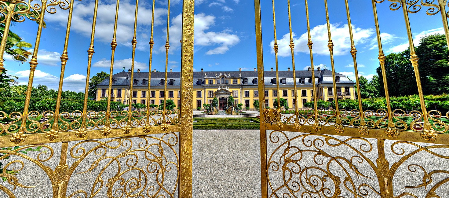 Blick durch ein prunkvoll verziertes, goldenes Eisengittertor auf das barocke Schloss Herrenhausen in Hannover. Im Vordergrund das geöffnete Tor mit floralen Ornamenten, im Hintergrund die symmetrische Gartenanlage mit Brunnen und gepflegten Beeten. Der Himmel ist strahlend blau mit einzelnen weißen Wolken – ein sonniger, klarer Tag.