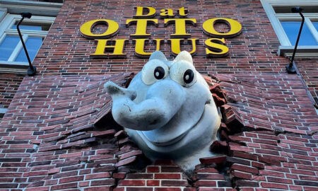 Außenfassade des Otto Huus in Emden mit auffälligem gelben Schriftzug „Dat OTTO HUUS“ auf rotem Klinker. Aus der Wand ragt ein großer grauer Ottifanten-Kopf, der scheinbar die Mauer durchbrochen hat. In den Fenstern links sind bunte Cartoonfiguren zu erkennen. Der Himmel ist teilweise bewölkt mit blauen Abschnitten.