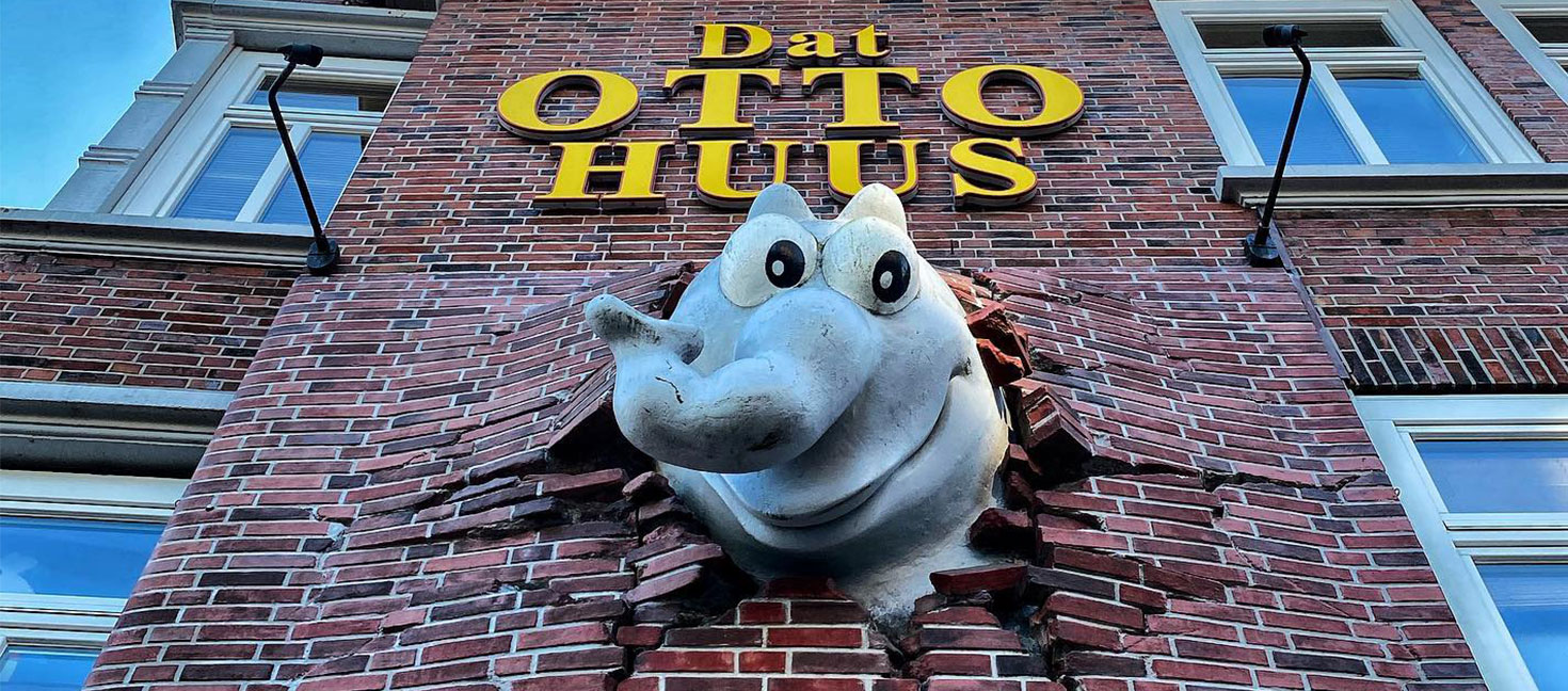 Außenfassade des Otto Huus in Emden mit auffälligem gelben Schriftzug „Dat OTTO HUUS“ auf rotem Klinker. Aus der Wand ragt ein großer grauer Ottifanten-Kopf, der scheinbar die Mauer durchbrochen hat. In den Fenstern links sind bunte Cartoonfiguren zu erkennen. Der Himmel ist teilweise bewölkt mit blauen Abschnitten.