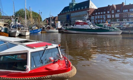 Ein kleines Ausflugsboot mit rotem Dach und dem Namen „SÜDREYERSHOF“ liegt am Rand eines breiten Kanals in Emden. Im Wasser schwimmen weitere Schiffe, darunter ein großes Segelboot. Im Hintergrund steht das grün-graue Rathaus mit Turm. Rechts sieht man Backsteinhäuser und blauen Himmel ohne Wolken.
