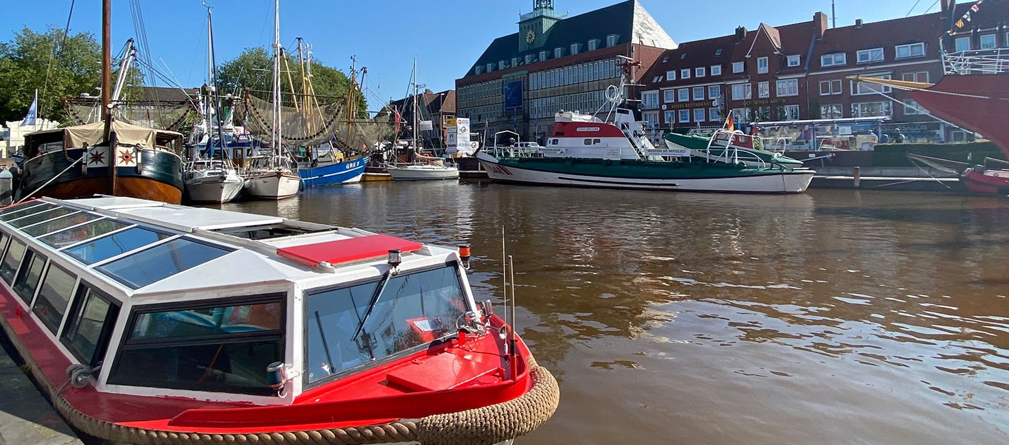 Ein kleines Ausflugsboot mit rotem Dach und dem Namen „SÜDREYERSHOF“ liegt am Rand eines breiten Kanals in Emden. Im Wasser schwimmen weitere Schiffe, darunter ein großes Segelboot. Im Hintergrund steht das grün-graue Rathaus mit Turm. Rechts sieht man Backsteinhäuser und blauen Himmel ohne Wolken.