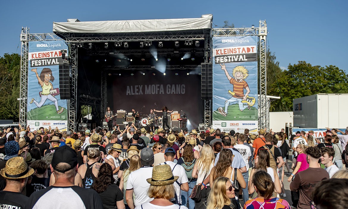 Ein Open-Air-Konzert beim „Kleinstadt Festival Meppen“. Auf einer großen Bühne tritt die Band „Alex Mofa Gang“ vor einer Menschenmenge auf. Viele Festivalbesucher*innen stehen dicht gedrängt, tragen Sommerkleidung und Strohhüte. Links und rechts der Bühne hängen große Banner mit Comicfiguren: links eine Frau mit Keyboard, rechts ein Junge mit E-Gitarre. Über beiden Figuren steht der Schriftzug „Kleinstadtfestival“. Im Hintergrund ist blauer Himmel und grünes Laub zu sehen – es herrscht sonniges Wetter.