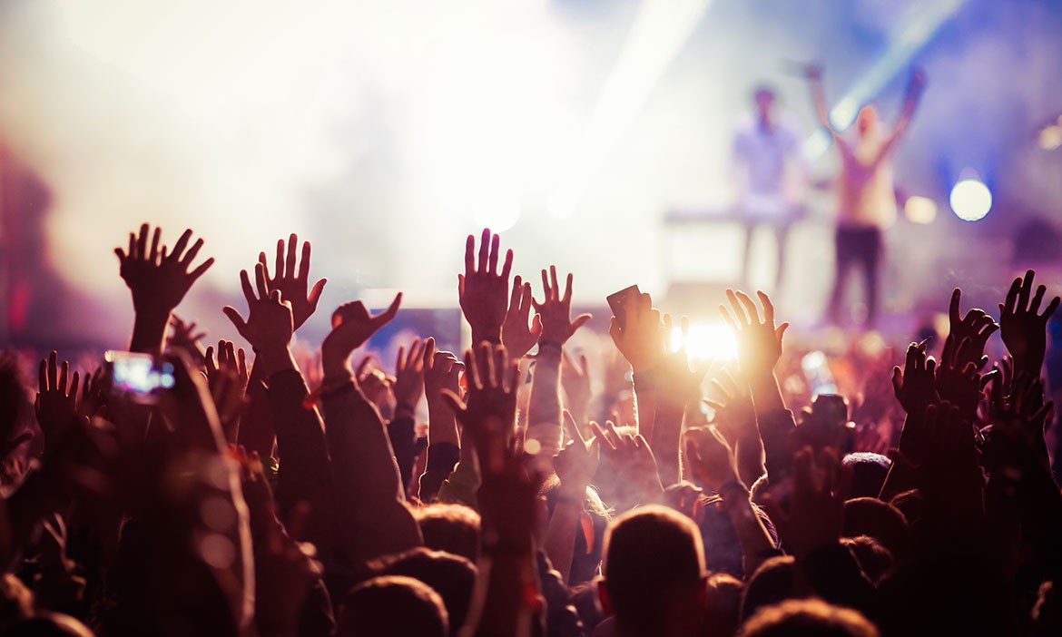 Menschenmenge bei einem Konzert oder Festival mit zahlreichen erhobenen Händen in der Luft. Im Vordergrund sind viele Besucher von hinten zu sehen, einige halten Smartphones hoch. Die Bühne im Hintergrund ist hell ausgeleuchtet, zwei Künstler sind nur schemenhaft durch das Gegenlicht und Nebel erkennbar. Die Stimmung wirkt energiegeladen, euphorisch und ausgelassen.