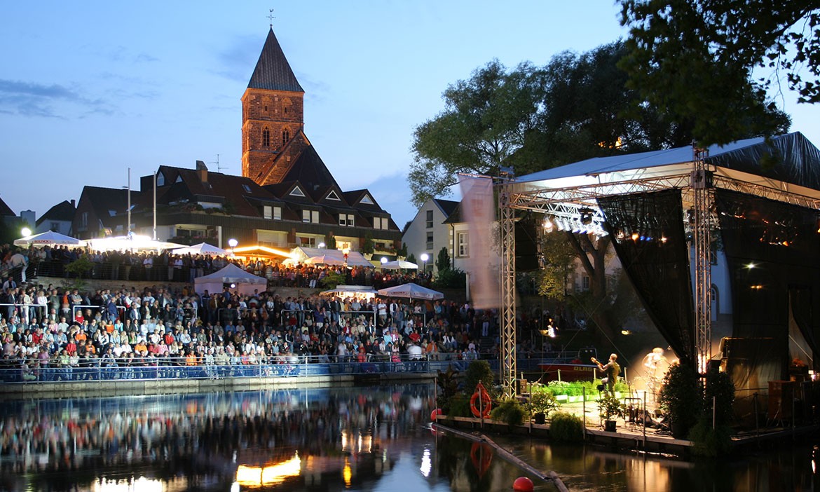 Abendstimmung beim EmsFestival in Rheine. Eine schwimmende Bühne ist auf dem Fluss aufgebaut und beleuchtet, darauf stehen Musiker und Musikerinnen im Scheinwerferlicht. Am gegenüberliegenden Ufer sitzt ein großes Publikum auf einer Tribüne und genießt das Konzert. Dahinter sind Fachwerkhäuser und eine Kirche mit beleuchtetem Turm zu sehen. Das Wasser spiegelt die Lichtstimmung der Bühne und der Stadt. Die Atmosphäre ist ruhig, festlich und sommerlich.