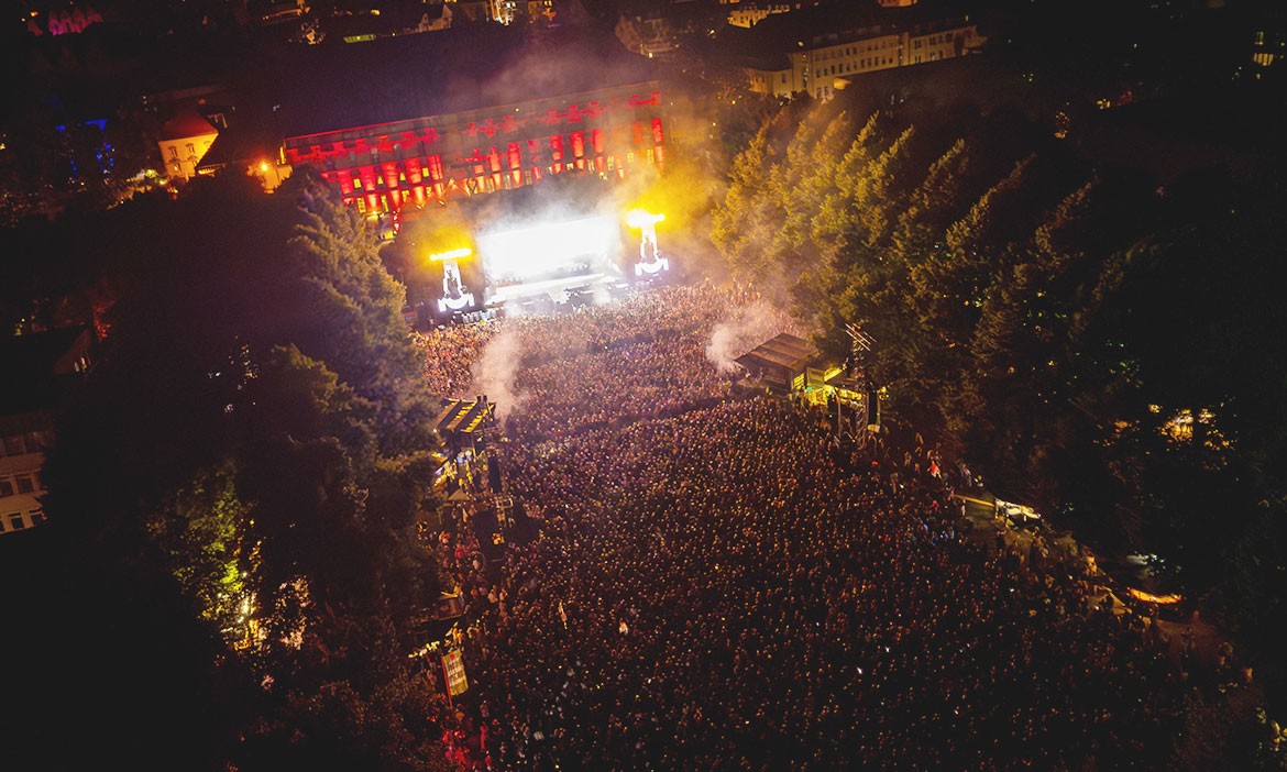 Luftaufnahme des Schlossgarten Open Airs in Osnabrück bei Nacht. Eine große Menschenmenge steht dicht gedrängt vor einer hell erleuchteten Bühne, von der Pyroeffekte und Lichtstrahlen in den dunklen Nachthimmel steigen. Im Hintergrund ist das historische Schloss in rotem Licht angestrahlt. Links und rechts der Bühne stehen große Bäume, die das Festivalgelände einrahmen. Rauchschwaden und die festliche Beleuchtung erzeugen eine eindrucksvolle Atmosphäre.